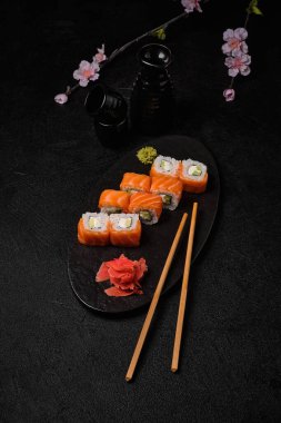 salmon sushi roll with red caviar, chopsticks, bottle of sake and cups on black concrete background