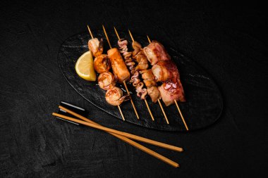 Japanese Seafood Skewers. Black tiger shrimp, scallop and salmon fillet, octopus served with lemon. Yakitori isolated on black concrete background. Pan Asian dish isolation. Kusiyaki seafood. Top view