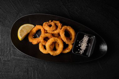 fried squid rings in breadcrumbs with lemon and soy sauce on black concrete background. Top view