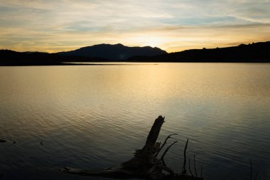 Beniarres rezervuarında gün batımında altın yansımalar ve arka planda Sierra de Mariola doğal parkı, İspanya