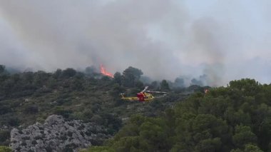 Tanımlanamayan bir itfaiye helikopteri, Yazı Tipi Roja Doğal Parkı yangınında, ateşin yanındaki tepenin arkasında kayboldu. Temmuz 2025, Ibi, İspanya.
