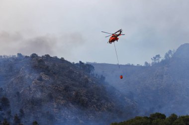 Temmuz 2025 'te Font Roja Doğal Park orman yangınında elinde Bambi kovası olan tanınmayan bir helikopter yangına yaklaşıyor.
