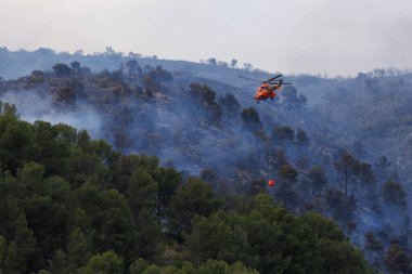 Temmuz 2025 'te Font Roja Doğal Park orman yangınında elinde Bambi kovası olan tanınmayan bir helikopter yangına yaklaşıyor.