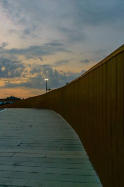 Hafif bulutlu bir gökyüzünde gün batımının arka planına doğru eğilen ahşap kenarlı bir köprü.