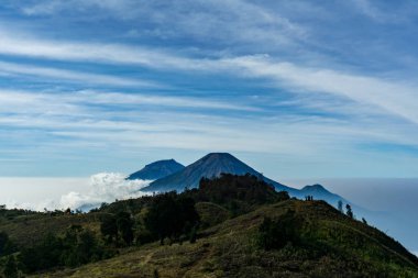 Sindoro Dağı, Sumbing ve Kembang 'ı pofuduk beyaz bulutlarla dolu canlı mavi bir gökyüzünün altında gösteriyor. Huzuru, korkuyu ve Endonezya 'nın volkanik manzaralarının güzelliğini aktarmak için mükemmel..