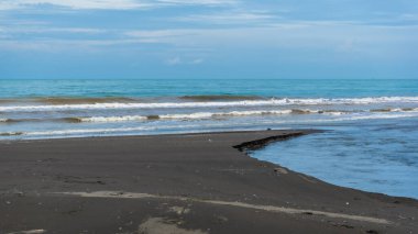 Derin mavi okyanus dalgalarıyla muhteşem Jodo Sahili beyaz bulutlu dramatik bir gökyüzünün altında siyah kuma çarpıyor.