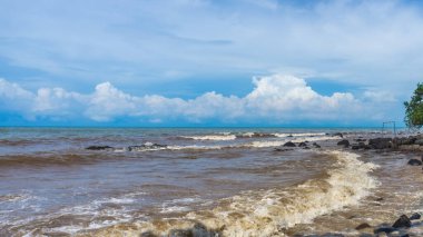 Endonezya, Batang 'da, Celong Sahili' ndeki kayalara çarpan güzel siyah-kahverengi dalgalar, beyaz bulutlu, çarpıcı mavi bir gökyüzünün altında.