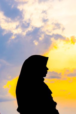 Tegal, Endonezya - 15 Eylül 2024: baş örtüsü içinde Asyalı Müslüman bir kadının silueti çarpıcı bir altın saat günbatımına karşı. Huzurlu ve güzel bir görüntü. Barış ve maneviyat için mükemmel.