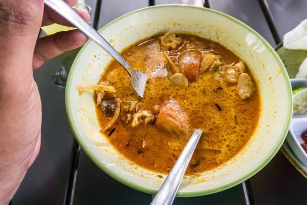 Bir insanın eli kaşığı tutuyor, lezzetli ve iştah açıcı Minapadi Körili Tavuk yemeğinin tadını çıkarıyor.