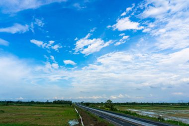 Yeşil tarlalardan geçen kamyon ve arabalı otoyol dramatik mavi gökyüzü ve bulutlar altında, ulaşım ve kırsal seyahat sahnesi