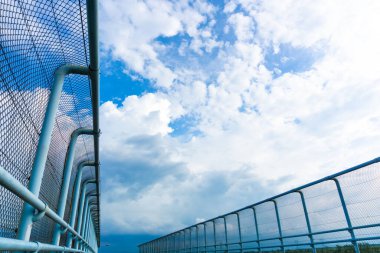 Mavi gökyüzüne ve dramatik bulutlara, kentsel altyapıya ve modern mimari tasarıma karşı perspektifli metal köprü çiti