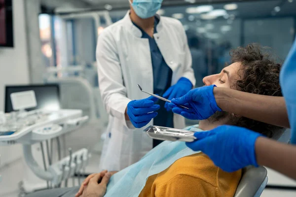 Dental assistant passing angled mirror dental instruments tools to a dentist with protective face mask and surgical gloves during patient's dental procedure standing in modern dentist's office.