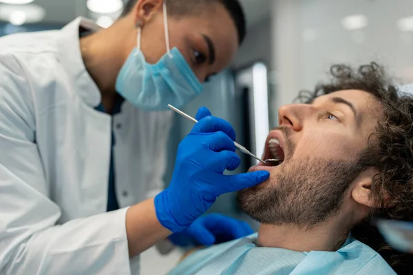 Young man with mouth open having dental exam during appointment at dentist\'s office, getting his teeth checked by female dentist with surgical mask and gloves at dental clinic.