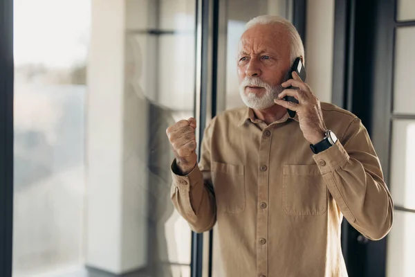 Angry senior businessman arguing over cell phone, feeling stressed, standing with raised fist near panoramic window in modern office showing urge to explain his point of view. Displeased old age man on phone.