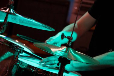 Siyah tişörtlü bir erkek müzisyenin elleri akustik davul çalıyor, ön planda ziller ve bagetler var, hepsi mavi ve yeşil renkte aydınlatılmış, arka planda odakları kesilmiş.