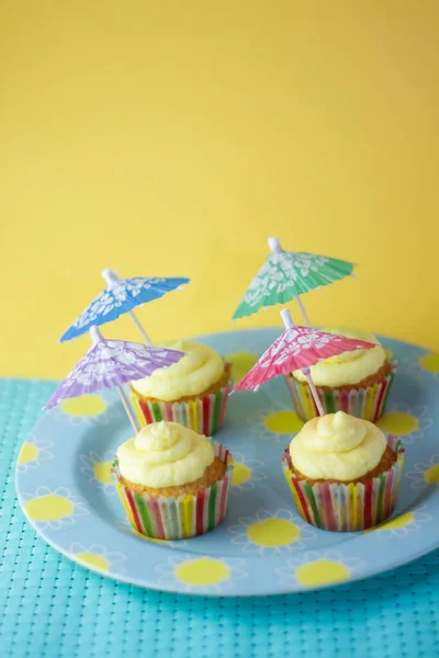 Four cupcakes with lemon cream and rainbow paper, with a cocktail umbrella, on top of a sky blue plate with drawn suns, on a sky blue tablecloth and a yellow background