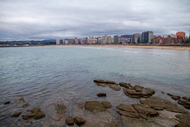 Gijon de Asturias şehrindeki San Lorenzo plajı, ön planda taşlar, arka planda binalar bulutlu bir günün gökyüzünü gösteriyor..