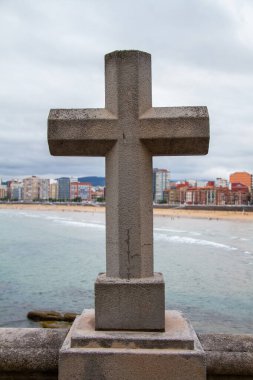 Gijon de Asturias şehrindeki San Lorenzo plajı, ön planda bir taş haç, arka planda ise bulutlu bir günün gökyüzünü gösteren binalar..