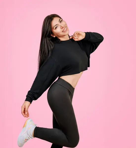 Young latina woman, nice body in black jumpsuit, posing cheerfully in studio with pink background.