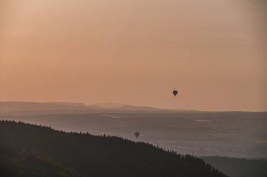 Renkli, canlı gökyüzüne karşı iki sıcak hava balonu. Arka planda resmedilmiş doğa manzarası.
