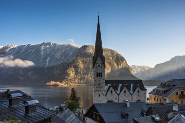Hallstatt, Avusturya manzarası, geleneksel mimarinin sergilendiği bir kilise kulesi, yemyeşil dağlar ve bulutlu bir gökyüzünün altında sakin bir göl manzarası.