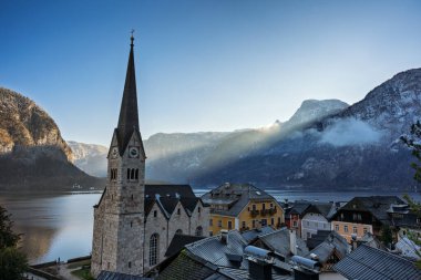 Hallstatt, Avusturya manzarası, geleneksel mimarinin sergilendiği bir kilise kulesi, yemyeşil dağlar ve bulutlu bir gökyüzünün altında sakin bir göl manzarası.