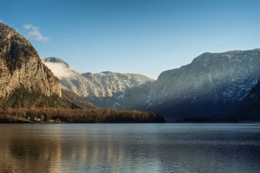 Hallstatt, Avusturya manzarası, geleneksel mimarinin sergilendiği bir kilise kulesi, yemyeşil dağlar ve bulutlu bir gökyüzünün altında sakin bir göl manzarası.