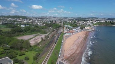 Goodrington Sands, Torbay, Güney Devon, İngiltere: DRONE AERIAL VİEWS: Buhar treni Paignton 'a doğru yolculuğuna devam ediyor; dalgalar güneşli bir bahar gününde Goodrington Kumları' na doğru ilerliyor.