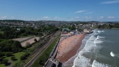 Goodrington Sands, Torbay, Güney Devon, İngiltere: DRONE VieWS: İHA, Goodrington Sands 'e doğru buharlı demiryolu hattına ve deniz kenarındaki dairelere doğru uçuyor; İHA, turistlerin güneşlenip denizde yüzerken deniz kıyısına doğru gelen dalgaları gösteriyor.