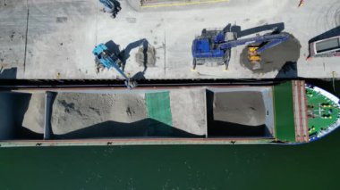 Teignmouth, Güney Devon, İngiltere: DRONE VİEWS: Bir kargo gemisi liman kenarı yükleyicileri tarafından top killeriyle doldurulur. Teignmouth limanı ayrıca hayvan yemi, toplamı ve tuz ithalatı ve ihracatıyla da ilgilenir; kasaba aynı zamanda popüler bir tatil beldesidir (Clip 4).