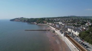 Dawlish, Güney Devon, İngiltere: DRONE VİEWS: Geriye doğru uçarak modern Dawlish tren istasyonu, demiryolu ve kumlu plajların üzerindeki deniz savunma duvarı üzerinden doğuya doğru uçar. Teignmouth hem bir liman hem de popüler bir İngiltere tatil beldesidir (Clip 2).