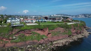 Hollicombe, Torquay, Torbay, Güney Devon, İngiltere: DRONE VİEWS: İHA yukarı ve yukarı pazar evleri ve körfezin karşısındaki Torquay kasabasını ortaya çıkarmak için kızıl kumtaşı uçurumları üzerinde yükselir. Torbay popüler bir İngiltere tatil beldesidir (Clip 2).