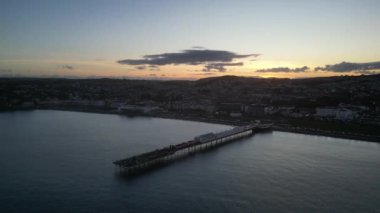 Paignton, Torbay, Güney Devon, İngiltere: DRONE VİEWS: Paignton Pier günbatımına karşı set. İskele, Kraliçe Victoria 'nın saltanatı sırasında 1879 yılında tamamlandı. Paignton, birçok turistik etkinliği olan popüler bir İngiltere tatil beldesidir (Clip 4).