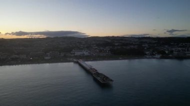 Paignton, Torbay, Güney Devon, İngiltere: DRONE VİEWS: Paignton Pier günbatımına karşı set. İskele, Kraliçe Victoria 'nın saltanatı sırasında 1879 yılında tamamlandı. Paignton, birçok turistik etkinliği olan popüler bir İngiltere tatil beldesidir (Clip 2).