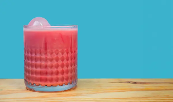 Sütlü pembe içecek, kristal bardakta soğuk içecek, ahşap masa üzerinde mavi arka planlı buz. Yaz, tatlı bir içki..