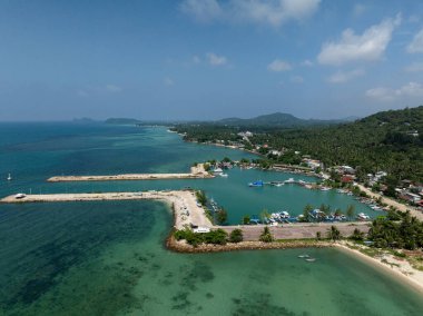 Açık mavi gökyüzünün altında, sahil şeridini çevreleyen tekneleri ve yemyeşil tepeleri olan geniş bir marina manzarası. Ko Pha Ngan, Tayland.