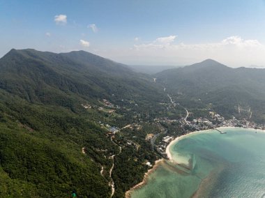 Yeşil yemyeşil tepeler ve turkuaz okyanus suları arasında uzanan küçük bir kasaba. Yollar ve plajlar görülebilir. Ko Pha Ngan, Tayland.