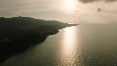 Güneşin batışı, okyanusa yansıyan altın ışık ve dramatik bulutlu bir gökyüzünün altındaki kıyı tepelerinin siluetleri. Ko Samui, Tayland.