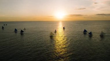 Yelkenliler, sakin tropikal bir ortamda gün batımının sıcak tonlarını yansıtan altın bir denizde hareket ederler. Boracay Adası, Filipinler.