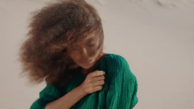 Kumlu çölde poz veren siyah elbiseli şık Afro-Amerikan model kız. Kıvırcık saçlı, tropik rüzgarlı çekici bir kadının portresi. Modern makyajlı hayalperest esmer kadın..