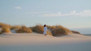Yalnız genç bir kadın yaz günü yüksek kuru çimenlerin önünde kum çölünde dikiliyor. Uzakta beyaz elbiseli kıvırcık Afrikalı Amerikalı kız vahşi doğada poz veriyor. Sakin esmer rüzgar saçlarını dalgalandırıyor..