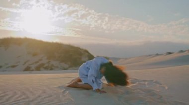 Tutkulu esnek kız dansçı gün batımında çölde serbest dans ediyor. Beyaz elbise giymiş, duygusal olarak kum atan Afro-Amerikalı genç bir kadın. Güzel anlamlı performans.