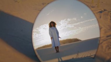Kum çölündeki aynada yansıyan şehvetli dans eden kadın. Genç Afro-Amerikalı kız beyaz elbise içinde el sıkışıyor. Kıvırcık esmer güzel günbatımı gökyüzünün önünde şarkı söylüyor.