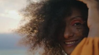 Modaya uygun kıvırcık kız kasvetli doğada kameraya gülümsüyor. Yaklaş. Afro-Amerikan mutlu kadın portresi. Rüzgarla savrulan gür saç modeline dokunuyor. Modern makyajlı güzel bir yüz..