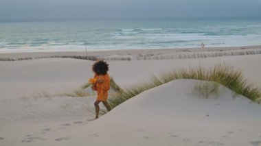 Yaz Alacakaranlığı 'nda boş kumsalda turuncu çiçeklerle yürüyen çekici genç bir kadın. Kıvırcık Afrikalı Amerikalı kız elinde güzel bir buketle yalınayak yürüyor. Kumral, deniz kıyısında rahatla.