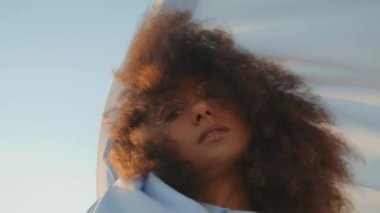 Şehvetli Afrikalı Amerikalı kız yaz mevsiminde gökyüzünün önünde çırpınan beyaz kumaşlarla dans ediyor. Çekici kıvırcık kadın, elinde bez tutarak doğada gösteri yapıyor. Baştan çıkarıcı esmer model kamera.