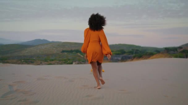 在夏夜黄昏时穿着橙色连衣裙在沙漠上慢跑的女子的背影 未知的卷曲女孩蹲伏在沙滩上 在漆黑的夜晚 活跃的非洲裔美国黑人在美丽的沙丘上放松一下 — 图库视频影像