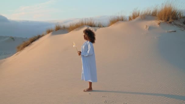 白い美しい花の夏の夜と砂の砂漠に立って穏やかなアフリカ系アメリカ人の女性 砂丘の荒野で1つの呼び出しを保持ポーズ白いドレスを着て魅力的なリラックスした女の子 自然概念 — ストック動画