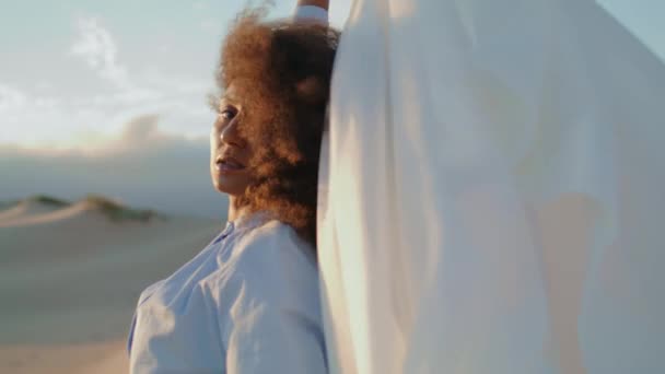砂の砂漠で夏の風を吹いて白い布を持つ誘惑的な深刻な女性が閉じます 若いアフリカ系アメリカ人の少女は 光の布のカメラを見てポーズ アートショー自然上のファッションパフォーマンス — ストック動画