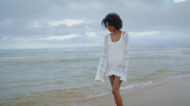 Beyaz kıyafetli, kumsala giden gülümseyen kadın. Çekici Afrikalı Amerikalı bulutlu bir günde okyanus dalgalarını izlemekten zevk alıyor. Güzel siyah saçlı kız sahil şeridinde rahatlıyor. Serene yaşam tarzı konsepti